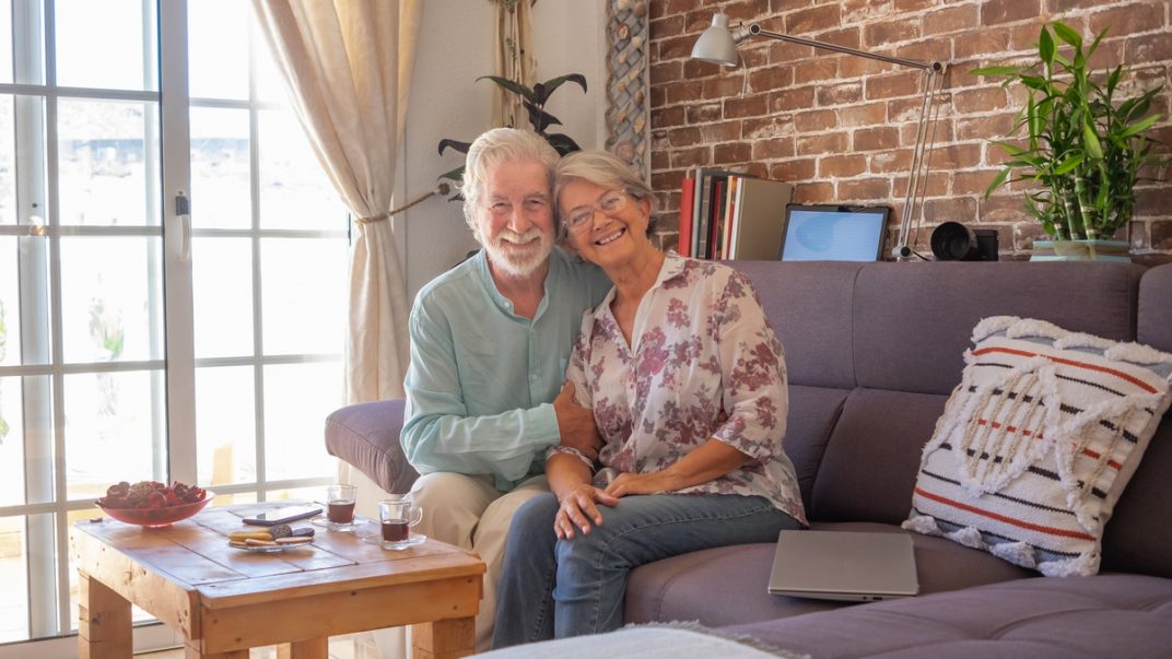 Continuing to Live at Home for Seniors, enjoying each other's company on a couch, with a view of the outside through a window.