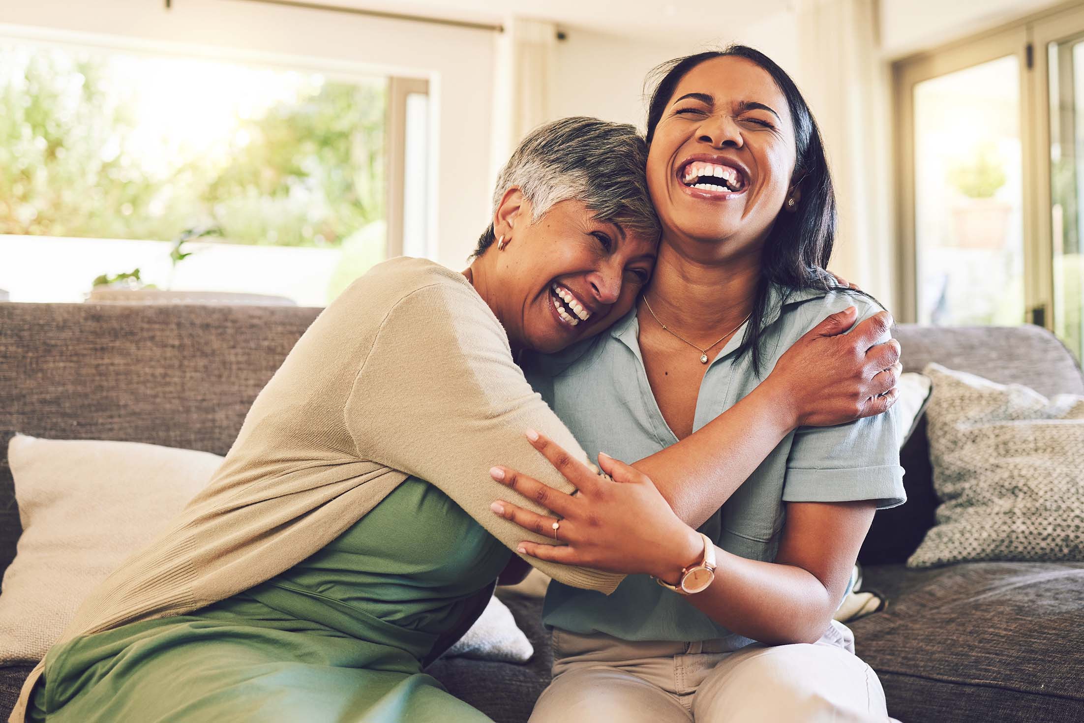 Two women share a joyful moment, laughing together on a couch, embodying peace of mind and friendship