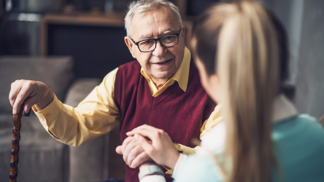 Tackling 3 common issues in people living with dementia with evidence-based tips 1 Caregiver holding the hand of an elderly man sitting on a couch with a cane.