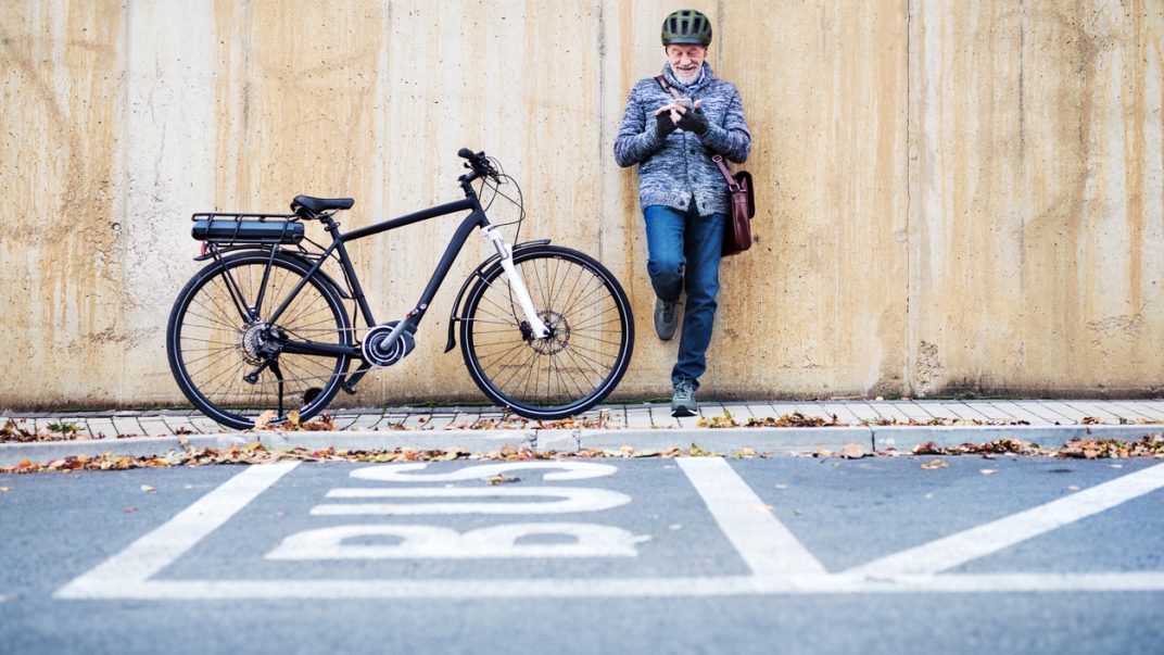 Alternatives to the car 1 Active senior man leaning against a concrete wall and using a smartphone with his bike next to him.
