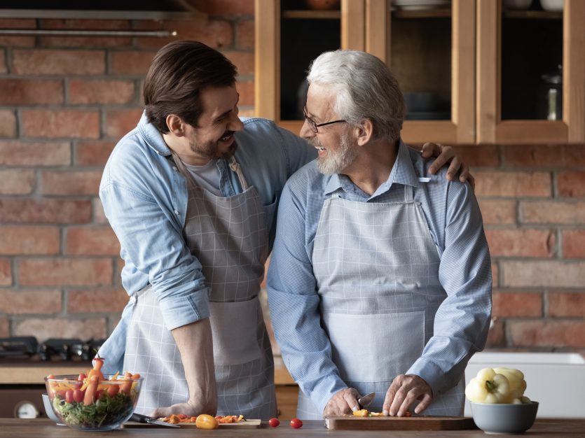 Elderly man and son cooking together, smiling.