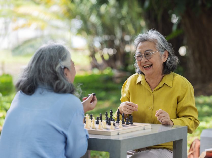Two older adults sitting in a park, playing chess, and laughing.