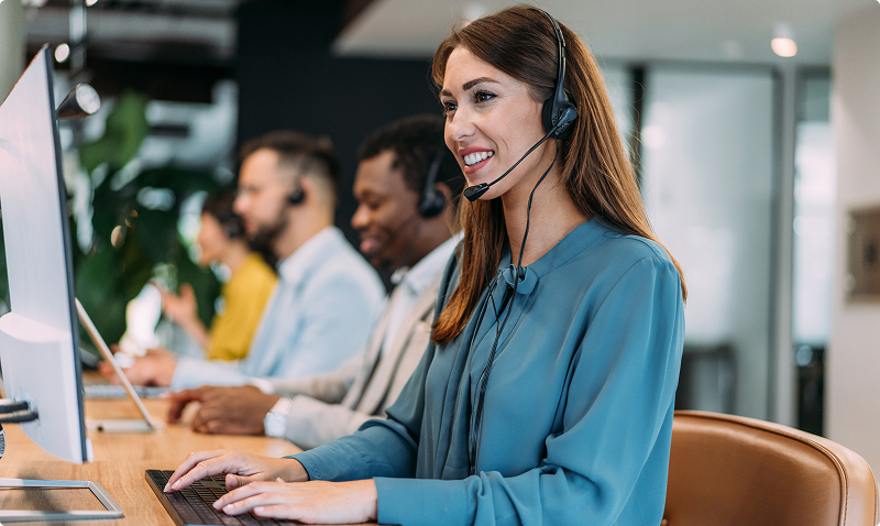 A woman with a headset sits at a desk in an office, focused on her work and engaged in a conversation.
